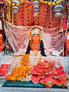 A vibrant and ornate statue of a deity adorned with a crown, surrounded by colorful fabric and garlands of marigold flowers. The background features an intricate, patterned orange and gold cloth. Offerings and religious items are placed in front, including flowers and a red booklet.