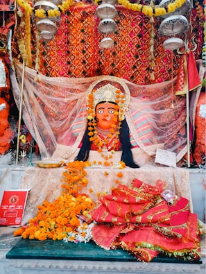 A vibrant and ornate statue of a deity adorned with a crown, surrounded by colorful fabric and garlands of marigold flowers. The background features an intricate, patterned orange and gold cloth. Offerings and religious items are placed in front, including flowers and a red booklet.