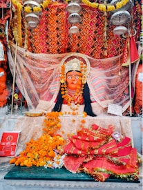 A vibrant and ornate statue of a deity adorned with a crown, surrounded by colorful fabric and garlands of marigold flowers. The background features an intricate, patterned orange and gold cloth. Offerings and religious items are placed in front, including flowers and a red booklet.
