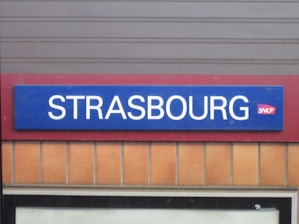 A rectangular train station sign with the word Strasbourg in white uppercase letters against a blue background. The SNCF logo is situated next to the text. The sign is mounted on a wall with a red border and beige tiles below.