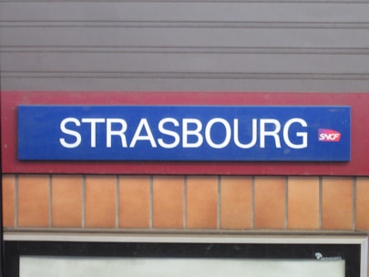 A rectangular train station sign with the word Strasbourg in white uppercase letters against a blue background. The SNCF logo is situated next to the text. The sign is mounted on a wall with a red border and beige tiles below.