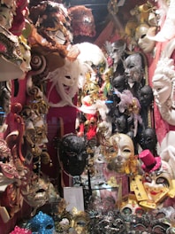 A collection of ornate and colorful Venetian masks displayed in a shop. The masks feature elaborate designs with feathers, gold detailing, and jewels. There are various styles, including full-face masks and eye masks, with some in vibrant colors like red, blue, and gold. A prominent black mask with intricate lace-like patterns is in the center.