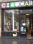 A pet store window display featuring various pet products such as scratching posts, pet food, and toys. The signage above the window has a playful design with colorful letters and a cat face within a letter. There is a poster on the door advertising a promotion. The weather appears overcast with some reflections on the wet pavement.