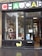 A pet store window display featuring various pet products such as scratching posts, pet food, and toys. The signage above the window has a playful design with colorful letters and a cat face within a letter. There is a poster on the door advertising a promotion. The weather appears overcast with some reflections on the wet pavement.