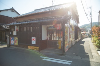 A traditional Japanese storefront with a tiled roof is located on a sunny street. The building is adorned with colorful posters and signs featuring Japanese characters and graphics. Adjacent to the shop, other buildings and a clear, lightly clouded sky are visible in the background, with mountains in the distance.