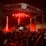 A large crowd gathers in front of a stage illuminated by bright red lights. The scene features a tall metal scaffolding and a DJ booth with electronic music equipment. Fog or smoke adds to the atmosphere as people raise their hands, capturing the vibrant energy of a live concert.