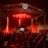 A large crowd gathers in front of a stage illuminated by bright red lights. The scene features a tall metal scaffolding and a DJ booth with electronic music equipment. Fog or smoke adds to the atmosphere as people raise their hands, capturing the vibrant energy of a live concert.