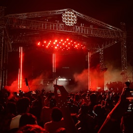 A large crowd gathers in front of a stage illuminated by bright red lights. The scene features a tall metal scaffolding and a DJ booth with electronic music equipment. Fog or smoke adds to the atmosphere as people raise their hands, capturing the vibrant energy of a live concert.