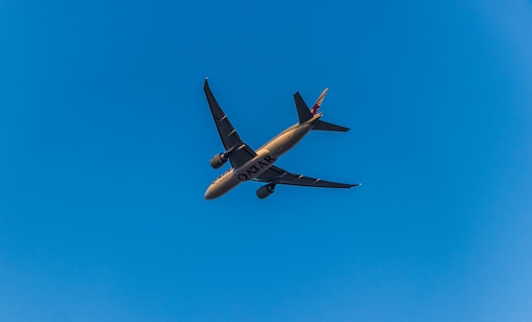 A commercial airplane flies against a clear blue sky, with the word 'Qatar' visible on the side of the aircraft. The airplane is captured from below, revealing the underside of its wings and fuselage.