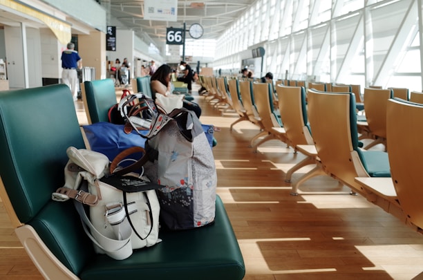 A spacious airport terminal features rows of green and wooden chairs with a few travelers seated throughout the area. In the foreground, a couple of bags, including a white backpack and a colorful tote, rest on the nearest chair. Natural light floods in through large windows on the right, illuminating the wooden floor and creating a modern, airy atmosphere.