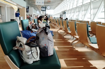 A spacious airport terminal features rows of green and wooden chairs with a few travelers seated throughout the area. In the foreground, a couple of bags, including a white backpack and a colorful tote, rest on the nearest chair. Natural light floods in through large windows on the right, illuminating the wooden floor and creating a modern, airy atmosphere.