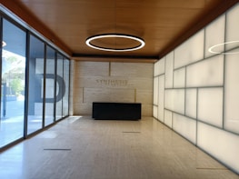 A modern interior space with a wooden ceiling and a circular light fixture. The walls are a combination of translucent and stone panels, creating a sleek and clean look. At the far end is a black rectangular reception desk with the word 'SYNTHESIS' engraved on the wall behind it. Large windows on the left allow natural light to flood the space, enhancing the minimalistic and open atmosphere.