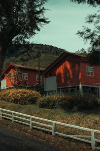 Two red wooden houses are set in a lush, green, hilly landscape. The foreground features a white wooden fence and a road running alongside manicured bushes and tall grasses. Trees surround the area, adding to the rural, serene atmosphere. Mountains are visible in the background under a clear sky, suggesting a peaceful countryside setting.