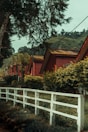 Several red-roofed wooden cabins are lined up along a path, surrounded by lush greenery and trees. A white wooden fence runs parallel to the row of cabins, delineating a boundary. In the background, there are hilly landscapes covered with trees.