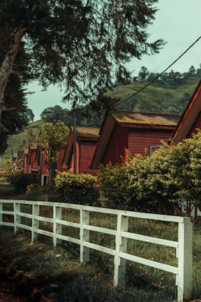 Several red-roofed wooden cabins are lined up along a path, surrounded by lush greenery and trees. A white wooden fence runs parallel to the row of cabins, delineating a boundary. In the background, there are hilly landscapes covered with trees.