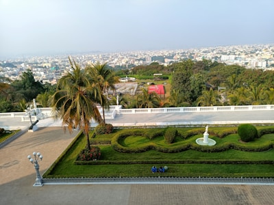 A well-maintained garden with ornate landscaping, including hedges, a palm tree, and a white statue at the center. In the foreground, there are two people sitting on the path, while the backdrop features a vast urban landscape with numerous buildings stretching to the horizon under a clear sky.
