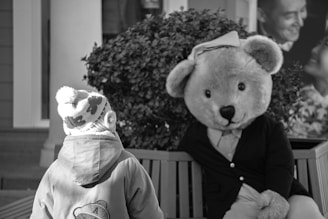 A child wearing a winter coat and a beanie with patterns stands facing a large teddy bear mascot seated on a bench. The teddy bear, dressed in a jacket and shirt, is positioned in a friendly manner. In the background, there's a bush and part of a poster showing a smiling couple.