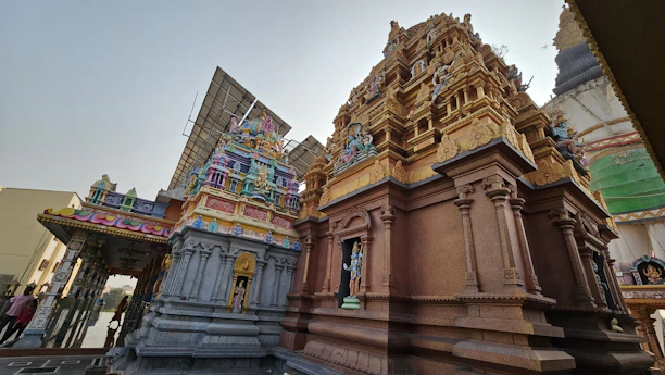 An intricately carved temple structure featuring vibrant statues and ornate designs, with solar panels visible in the background. The temple showcases a combination of colorful deities and traditional architectural elements.