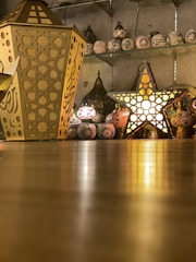 The image features an indoor display showcasing a variety of ornamental lanterns and decorative lamps made of metal and glass. Prominently placed on the floor are a large, hexagonal lantern with intricate cut-out patterns and a star-shaped lantern with luminous light emanating through its perforations. In the background, several other lanterns are arranged on shelves against a wall, contributing to a warm and inviting ambiance.