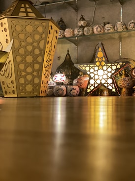 The image features an indoor display showcasing a variety of ornamental lanterns and decorative lamps made of metal and glass. Prominently placed on the floor are a large, hexagonal lantern with intricate cut-out patterns and a star-shaped lantern with luminous light emanating through its perforations. In the background, several other lanterns are arranged on shelves against a wall, contributing to a warm and inviting ambiance.