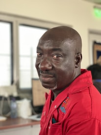 A man wearing a red uniform with embroidered text is seated indoors, possibly in an office or workspace. The background features blurred elements including a window, a computer monitor, and other desk items.
