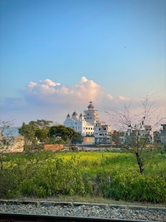 A serene landscape featuring a large, intricate temple with multiple spires in the distance. The setting includes lush green fields and sparse trees in the foreground, with a railway track running parallel. The sky is clear with a few fluffy clouds, indicating calm weather.