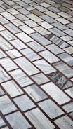 A detailed pattern of rectangular marble tiles with varying shades of gray and white, bordered by thin dark brown grout lines. The tiles are laid out in an offset arrangement, creating a visually interesting grid.