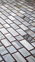 A detailed pattern of rectangular marble tiles with varying shades of gray and white, bordered by thin dark brown grout lines. The tiles are laid out in an offset arrangement, creating a visually interesting grid.