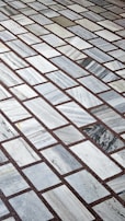 A detailed pattern of rectangular marble tiles with varying shades of gray and white, bordered by thin dark brown grout lines. The tiles are laid out in an offset arrangement, creating a visually interesting grid.