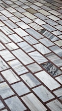 A detailed pattern of rectangular marble tiles with varying shades of gray and white, bordered by thin dark brown grout lines. The tiles are laid out in an offset arrangement, creating a visually interesting grid.