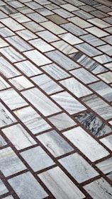 A detailed pattern of rectangular marble tiles with varying shades of gray and white, bordered by thin dark brown grout lines. The tiles are laid out in an offset arrangement, creating a visually interesting grid.