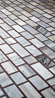 A detailed pattern of rectangular marble tiles with varying shades of gray and white, bordered by thin dark brown grout lines. The tiles are laid out in an offset arrangement, creating a visually interesting grid.