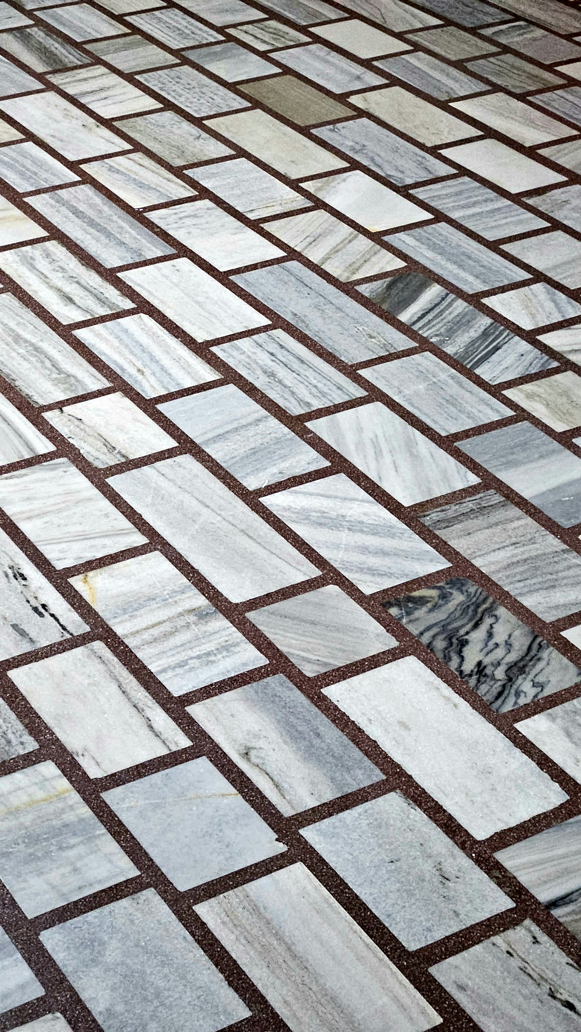 A detailed pattern of rectangular marble tiles with varying shades of gray and white, bordered by thin dark brown grout lines. The tiles are laid out in an offset arrangement, creating a visually interesting grid.