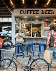 A small outdoor café serves coffee and beer. People are seen interacting with the establishment, including a person sitting on a blue stool with their back turned and another holding a shopping bag. A bicycle is parked in the foreground, and the café's countertop displays several beverages.