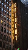 An exterior view of a building featuring a large vertical sign with illuminated yellow letters spelling 'COLBERT', set against a backdrop of windows and scaffolding.