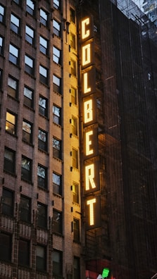 An exterior view of a building featuring a large vertical sign with illuminated yellow letters spelling 'COLBERT', set against a backdrop of windows and scaffolding.