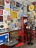 A wall covered with a diverse collection of vintage and retro-style signs and posters. The area features some circular and rectangular tables with red chairs. There are advertisements and travel-related signage including brands like Red Bull and Pepsi, and references to locations such as New York and Alabama. A prominent yellow and black circular sign with a telephone symbol is also visible.
