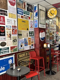 A wall covered with a diverse collection of vintage and retro-style signs and posters. The area features some circular and rectangular tables with red chairs. There are advertisements and travel-related signage including brands like Red Bull and Pepsi, and references to locations such as New York and Alabama. A prominent yellow and black circular sign with a telephone symbol is also visible.