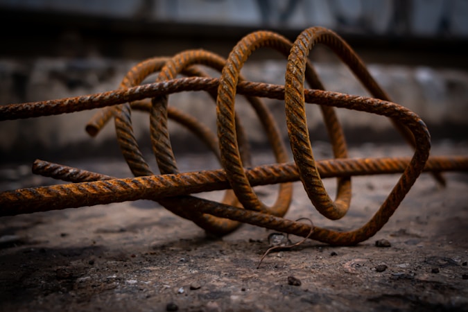 Coiled rusted metal rods rest on a rough, rocky surface. The texture of the metal is gritty with visible signs of corrosion. The background is blurred, emphasizing the twisted, industrial shapes in the foreground.