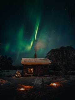 A wooden cabin with snow on the roof sits in the midst of a snowy landscape. Warm light emanates from the window, creating a cozy atmosphere. Above, the green and turquoise hues of the Northern Lights illuminate the night sky, casting a magical glow.