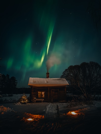 A wooden cabin with snow on the roof sits in the midst of a snowy landscape. Warm light emanates from the window, creating a cozy atmosphere. Above, the green and turquoise hues of the Northern Lights illuminate the night sky, casting a magical glow.