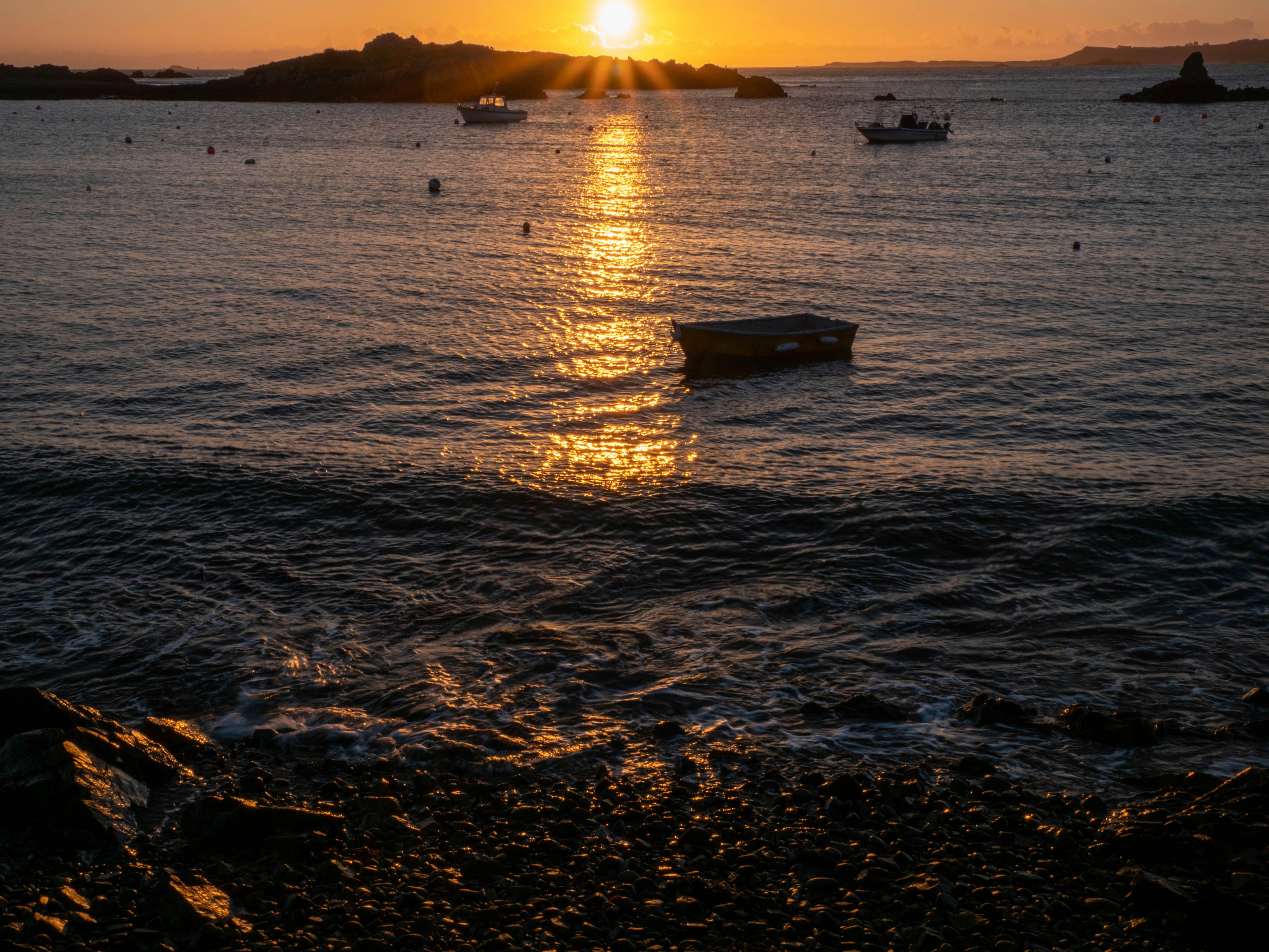 a boat floating on top of a body of water, A sunrise at Bordeaux Beach, Guernsey with Herm in the background.