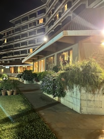 A multi-story building with illuminated windows at night, featuring a modern architectural design. Lush greenery and potted plants line the walkway leading to the entrance, with well-maintained lawns and foliage providing a serene atmosphere. Soft lighting enhances the ambiance, creating a welcoming environment.