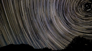 A night sky photograph featuring long exposure star trails creating circular patterns, with a silhouette of a landscape at the bottom.