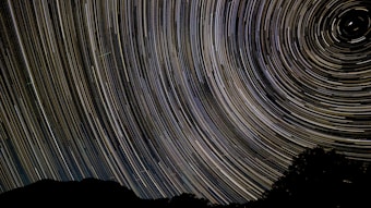 A night sky photograph featuring long exposure star trails creating circular patterns, with a silhouette of a landscape at the bottom.