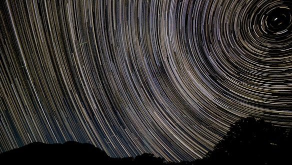 A night sky photograph featuring long exposure star trails creating circular patterns, with a silhouette of a landscape at the bottom.