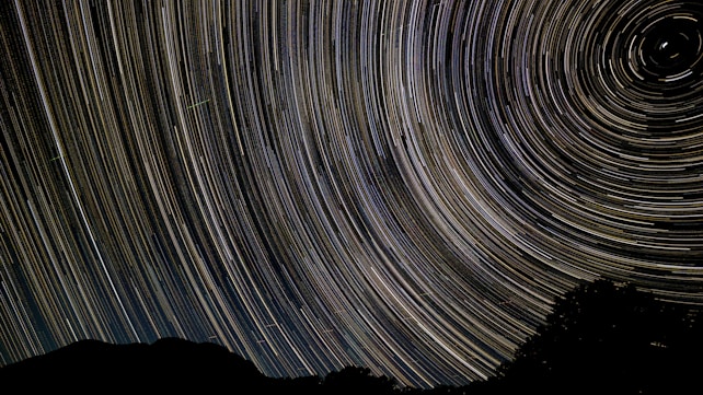 A night sky photograph featuring long exposure star trails creating circular patterns, with a silhouette of a landscape at the bottom.