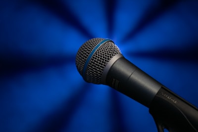 A close-up view of a handheld microphone with a textured metal grille and a dark matte finish, set against a vibrant blue background that radiates soft light effects.