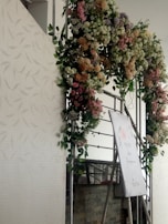 A decorative floral arrangement displayed above an easel. The arrangement features a variety of flowers in different colors, including pastel shades of pink, white, and purple. The easel holds a sign or poster with some text, partially visible, amidst the flowers. It appears to be an event setup with a delicate and floral theme.