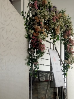 A decorative floral arrangement displayed above an easel. The arrangement features a variety of flowers in different colors, including pastel shades of pink, white, and purple. The easel holds a sign or poster with some text, partially visible, amidst the flowers. It appears to be an event setup with a delicate and floral theme.